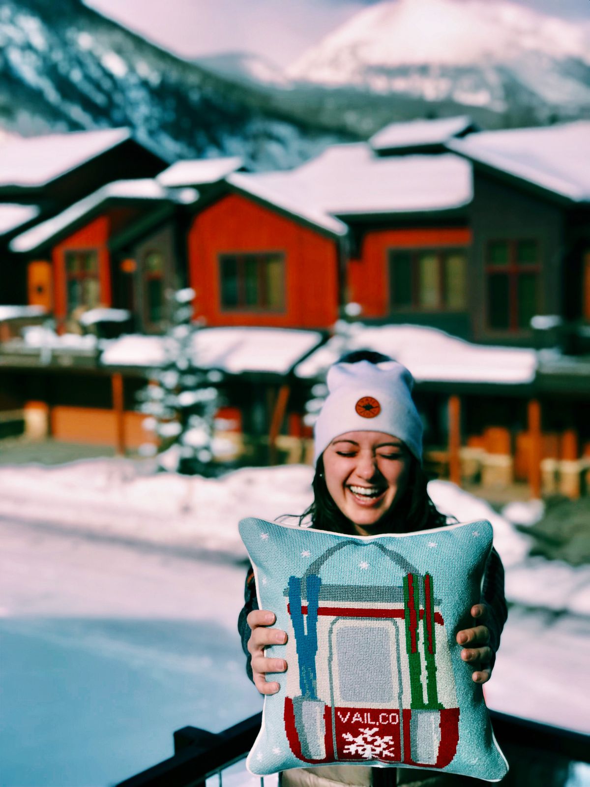 Woman laughing while holding a Vail needlepoint pillow on a snowy village street
